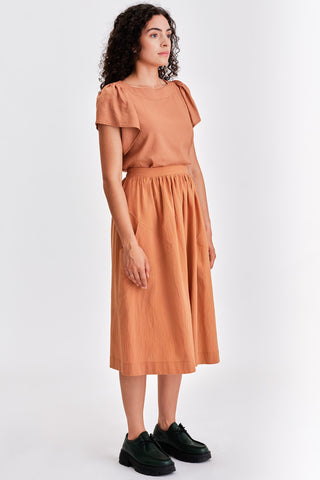 Woman in front of white background wearing caramel orange blouse, matching voluminous midi length skirt and black leather lace-up shoes. 