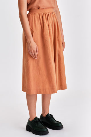 Woman in front of white background wearing caramel orange voluminous midi length skirt and green leather lace-up shoes. 