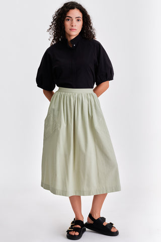 Woman in front of white background wearing black blouse, voluminous light green midi length skirt and black leather sandals. 