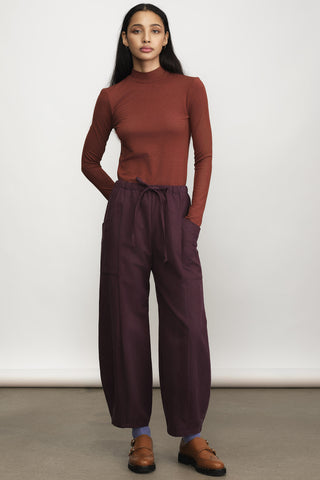 A woman wearing a rust mock neck long-sleeve top and dark plum  drawstring waist pants, paired with brown leather loafers, against a neutral studio backdrop.