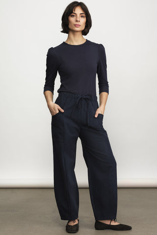 A woman wearing  a black long sleeve top and black drawstring waist pants with brown flats, against a neutral studio background. 