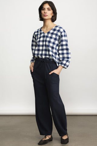 A woman wearing a blue and white check long sleeve  top paired with navy drawstring waist and black flats, standing against a plain light gray studio background.