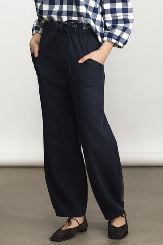 A woman wearing a blue and white check long sleeve  top paired with navy drawstring waist and brown flats, standing against a plain light gray studio background.
