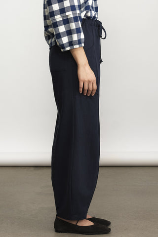A side-profile view of a person wearing dark navy wide-leg drawstring pants, a blue and white checkered shirt, and brown flats against a plain studio background.