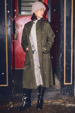 Woman standing outside in front of red door wearing an olive below the knee coat with beige toque, cream dress and black leather biker boots. 