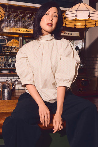 Woman sitting on bar top in bar waring cream coloured mock neck top with pleated half sleeves paired with black pants. 