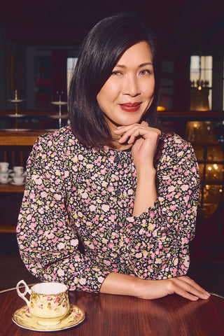 Woman sitting at a table with teacup, wearing black floral button up shirt. 