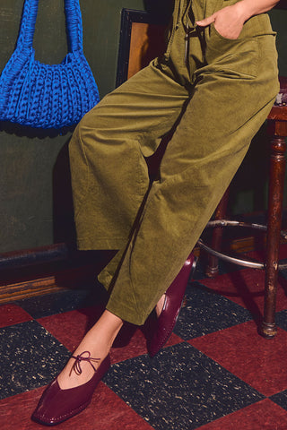 Woman sitting on bar stool, wearing olive pants, a blue crocheted bag and burgundy patent leather tie up ballet flats. 