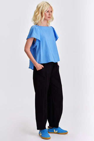 Woman wearing blue blouse, black pants and blue sneakers in front of white background. 