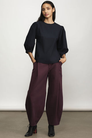 A model wearing a black long-sleeve top with voluminous sleeves and burgundy barrel-cut pants, paired with black lace-up ankle boots, against a neutral studio backdrop.