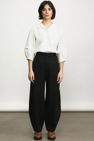 A model stands against a plain light gray studio wall wearing a white long-sleeve button-up shirt tucked into high-waisted black barrel-cut trousers, with brown shoes.