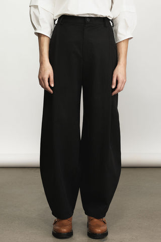 A person wearing black barrel cut trousers with a white long-sleeve top and brown shoes, standing against a plain light gray studio background.