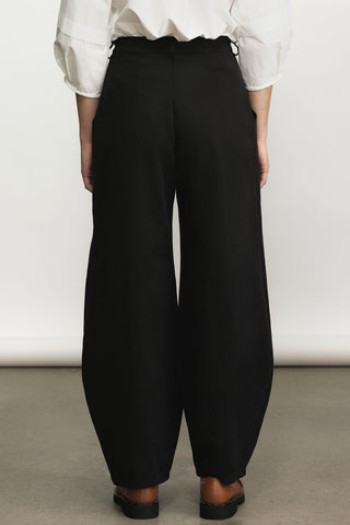 A person shown from the back wearing black barrel-cut trousers with a white blouse and black shoes, standing against a plain light wall.