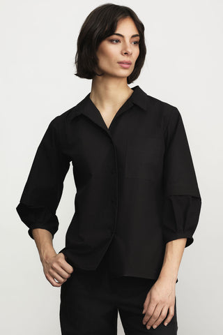 A woman modeling a black button-up shirt with three-quarter sleeves, paired with black pants, against a light gray studio background.