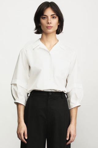 A woman modeling a white button-up shirt with three-quarter sleeves, paired with black pants, against a light gray studio background.