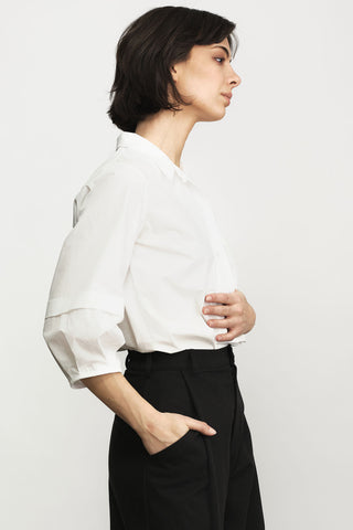 A woman in side profile wearing a white long-sleeve button-up shirt tucked into black trousers, photographed against a clean light gray studio background.