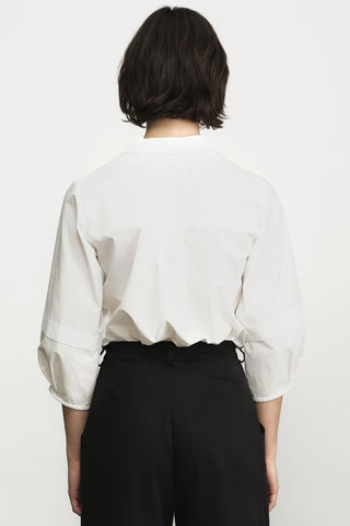 A person shown from the back wearing a white long-sleeve blouse tucked into black trousers, against a plain light background.