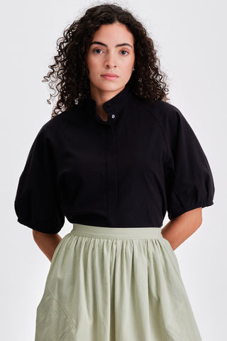 Woman in front of white background wearing black blouse with voluminous sleeves tucked into light green skirt. 