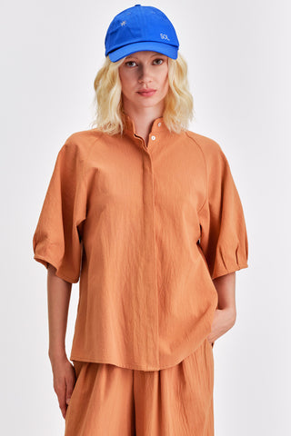 Woman in front of white background wearing bright blue hat, caramel blouse with voluminous sleeves and matching pants. 