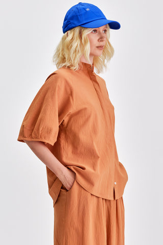 Woman in front of white background wearing bright blue hat, caramel blouse with voluminous sleeves and matching pants. 