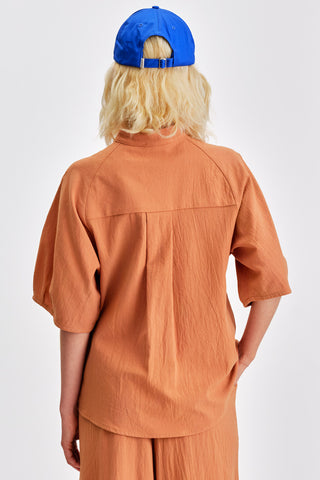 Back view of woman in front of white background wearing bright blue hat, caramel blouse with voluminous sleeves and matching pants. 