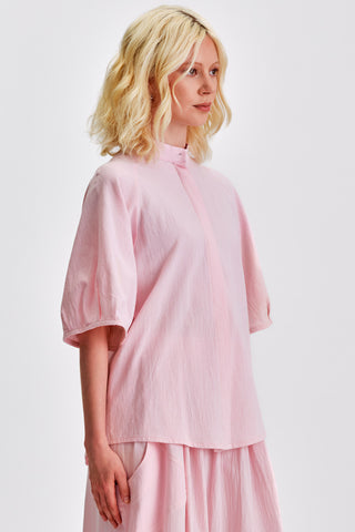 Woman in front of white background wearing pink blouse with voluminous sleeves and matching skirt. 