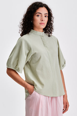 Woman in front of white background wearing light green blouse with voluminous sleeves. 