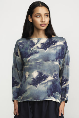 A woman wearing a long-sleeve boat-neck top with a blue and gray watercolor cloud pattern, paired with dark navy pants, against a plain light gray background.