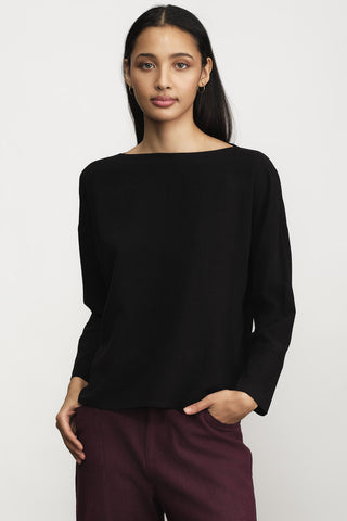 A woman modeling a black long-sleeve boat-neck top with dark burgundy pants, standing against a light gray studio background.