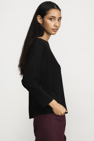 A woman wearing a black long-sleeve top and burgundy pants, shown in profile against a plain light gray studio background.