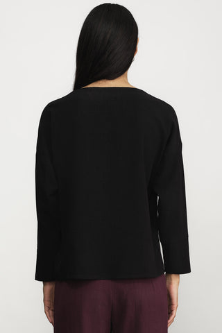A woman viewed from the back wearing a black long-sleeve top and dark burgundy pants against a plain light gray studio background.