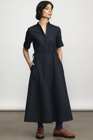 Model in front of white wall wearing navy blue midi length dress with button front and tie at waist. 