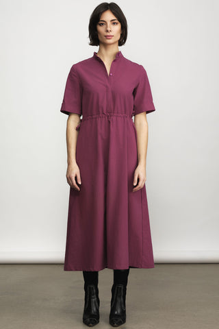 Model in front of white wall wearing light burgundy midi length dress with button front and tie at waist. 