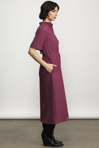 Side view of model in front of white wall wearing light burgundy midi length dress with button front and tie at waist. 