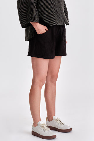 Woman in front of white background wearing black mid-thigh length shorts with pockets and white sneakers. 