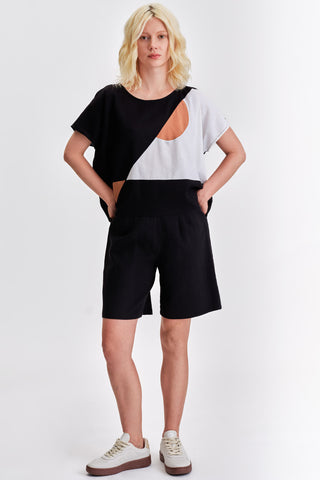 Woman in front of white background wearing black and white colour blocked shirt, black pleated bermuda shorts and white sneakers. 