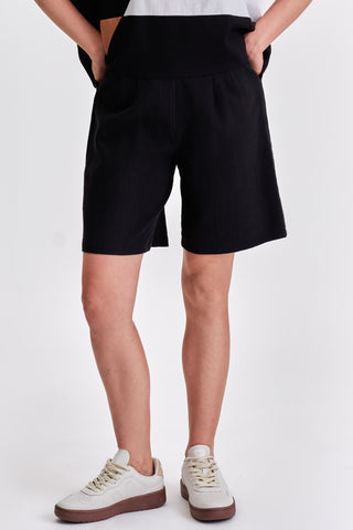 Woman in front of white background wearing black pleated bermuda shorts and white sneakers. 