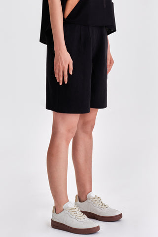 Side view of woman in front of white background wearing black pleated bermuda shorts and white sneakers. 