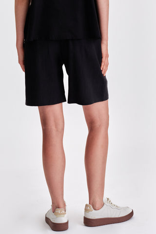 Back view of woman in front of white background wearing black bermuda shorts and white sneakers. 