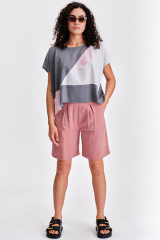 Woman in front of white background wearing grey and white colour blocked shirt and pink pleated bermuda shorts. 