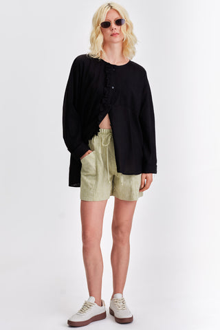 Woman in front of white background wearing black button up shirt and light green mid-thigh shorts with front pockets. 