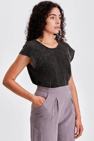 Woman in front of white background wearing black and white stripe cap sleeve top and grey pants. 