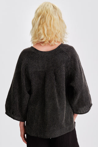 Back view of woman in front of white background wearing black striped loose fitting blouse with voluminous sleeves. 