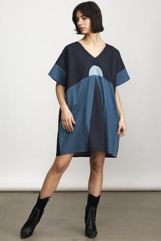 A woman wearing a loose, short-sleeve V-neck dress with a geometric color-blocked pattern in navy, medium blue, and light blue, paired with black ankle boots against a neutral studio background.