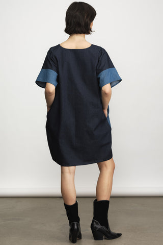 A model viewed from behind wearing a loose-fitting navy blue dress with contrasting medium-blue denim sleeve cuffs, paired with black ankle boots, against a plain light gray studio background.