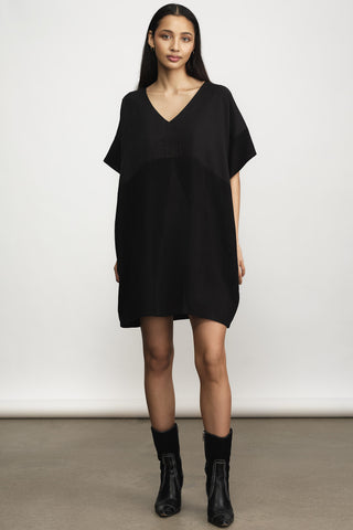 A woman modeling a loose-fitting black V-neck mini dress with short sleeves, paired with black ankle boots, against a plain light gray studio background.