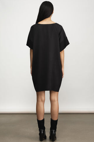 A woman viewed from behind wearing an oversized black short-sleeve dress with black socks and black heels, standing against a plain light gray studio wall.