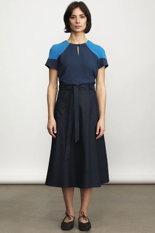 A woman stands against a plain light gray studio wall wearing a color-block midi blouse with a navy a-line skirt, paired with dark flats.