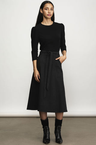 A woman modeling a black three-quarter sleeve top and a black midi a-line skirt with a tie waist, paired with black socks and black ankle boots, against a neutral studio background.