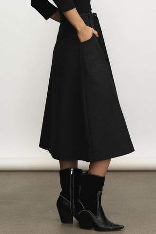 A person wearing a black midi a-line skirt with side pockets and black ankle boots, standing against a plain light gray studio background.